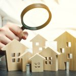 a person holding a magnifying glass over a wooden houses. Real estate appraiser. Property valuation / appraisal. Find a house. Search for housing. Real estate market analysis. Selective focus